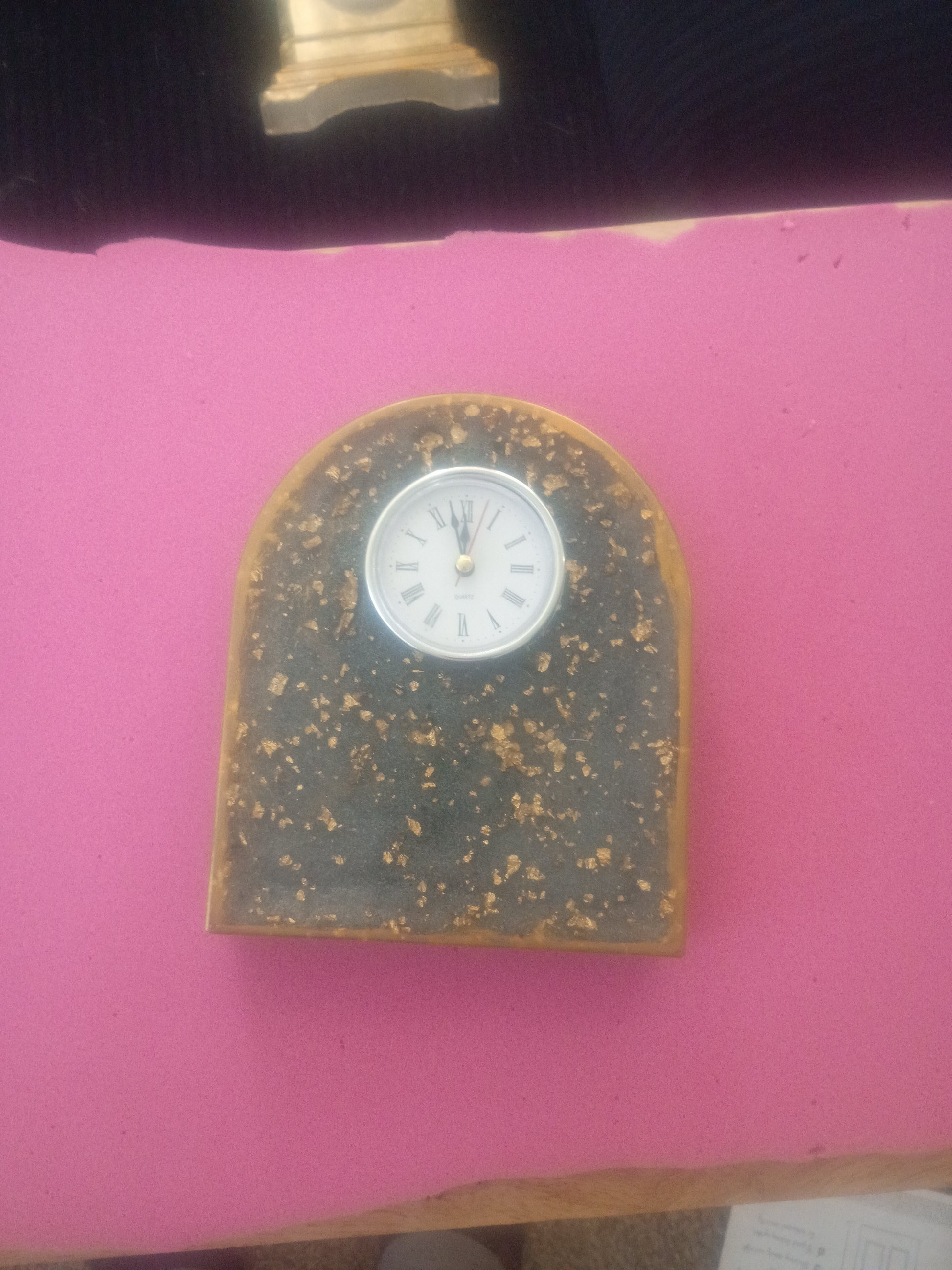 A small arched tabletop clock with a speckled dark gray or black stone-like face, a gold-toned border, and a white circular dial, photographed on a bright pink surface.